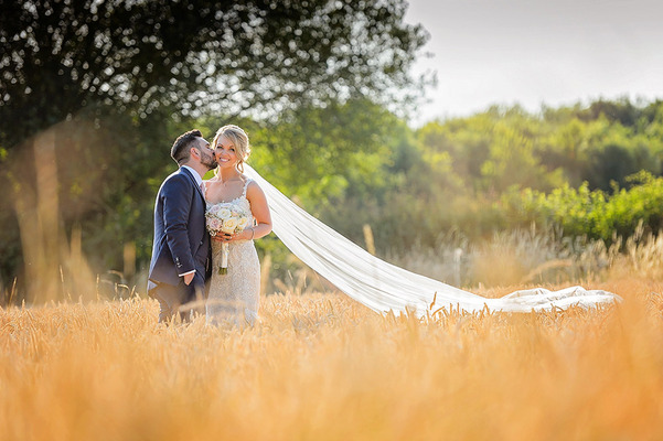 Groom kissing bride with long veil on the cheek  in field